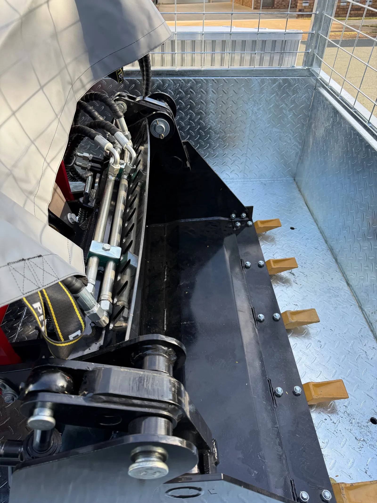 Black toothed bucket and hydraulic hoses on the Rhinoceros mini loader, inside a galvanised checker-plate cage trailer