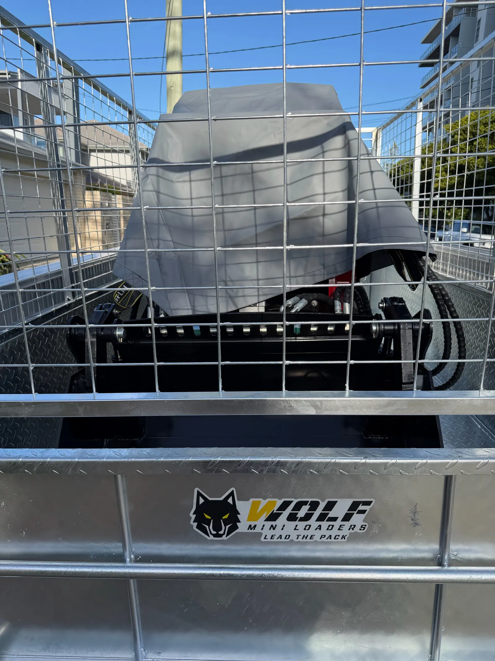 Mini loader under a grey cover, loaded in a galvanised cage trailer, Wolf Mini Loaders Lead the Pack sticker on the trailer front